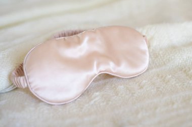 Apricot pink satin eye sleeping mask on beige knitted background. Insomnia and sleeplessness disorder concept. Close up, copy space, flat lay. Accessories for girls and women