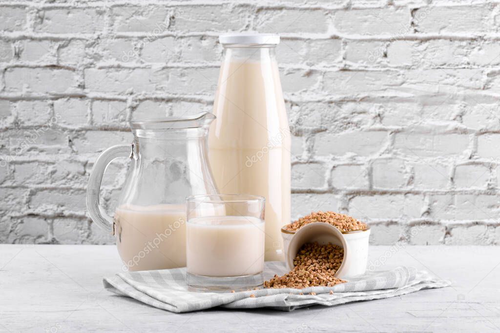 Vegan non diary buckwheat milk in bottle, in jug and glass with buckwheat groats in a bowl on white stone table. Vegan buckwheat drink is plant based alternative milk. Veggies healthy milk product
