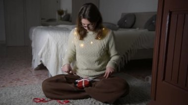 Gözlüklü, bej süveter giyen, Noel 'i, doğum gününü ya da sevgililer gününü kutlayan kırmızı hediyeli ve yatak odasının yanındaki halının üzerinde oturan 4k kızı. Tatil ve satış sezonu için hazırlık