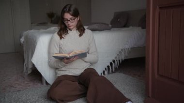 4k gözlüklü, süveter giyen genç bir kadın, sıcacık sarılma odasında otla kitap okuyor, sayfayı çeviriyor, öğreniyor, çalışıyor, eğitim, rahatlama. Evde sıcak bir kış günü..