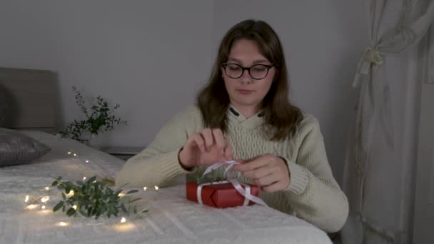 4K中国英语学习网一个戴眼镜 头戴米黄色毛衣的女孩在晚上 寒假和销售旺季的床上用红色牛皮纸包礼物 — 图库视频影像