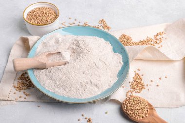 Mavi seramik tabakta karabuğday unu, ahşap kepçe kepçe, kabın üzerinde çiğ yeşil karabuğday tohumu ve açık gri masa arkasında peçete. Alternatif un, glütensiz un, sağlıklı beslenme.