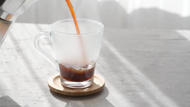 新鮮なコーヒーは透明なコーヒーカップにモカから注いでいます その隣には 灰色のテーブルの背景に窓から朝の影と朝の日差しがあるチョコレートチップクッキーがあります — ストック動画