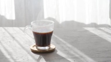 Sabah gölgelerinde sıcak kahve fincanı ve gri masa arkasındaki pencereden güneş ışınları. Gri masa arkasında buharlı, şeffaf cam kahve fincanı.