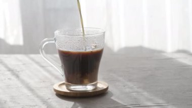 Süt, şeffaf bir kahve fincanında taze bir kahveye dökülüyor. Sabah gölgeleri ve sabah güneşleri gri bir masa arkaplanındaki pencereden içeri akıyor.