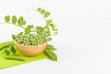 Tahta bir kasede taze yeşil bezelyeler ve bezelye bitkileri beyaz arka planda yeşil bir peçete üzerinde yeşil yapraklar, sağlıklı fasulye proteinleri.