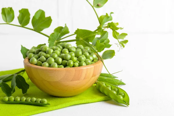 Ervilhas verdes frescas em uma tigela de madeira com plantas de ervilhas folhas em um guardanapo verde no fundo branco, proteína de feijão saudável — Fotografia de Stock