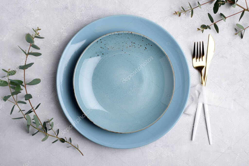 Festive table setting with blue ceramic plate, golden cutlery set and fresh eucalyptus leaves on grey concrete table top view. Plate mockup. Copy space, element for design