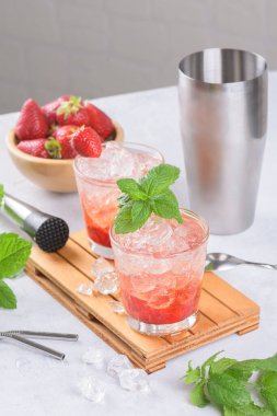 Soğuk yaz çilekli mojito, margarita, daiquiri. İki bardak taze çilekli soda, buz küpleri, nane şekeri ve açık arkaplanda barmen aletleri. Buzlu çilekli limonata, yakın çekim.
