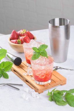 Soğuk yaz çilekli mojito, margarita, daiquiri. İki bardak taze çilekli soda, buz küpleri, nane şekeri ve açık arkaplanda barmen aletleri. Buzlu çilekli limonata, yakın çekim.