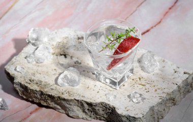 Vodka in shot glass with ice, decorated with strawberry slice and thyme on travertine podium on pink marble stone background. Alcohol cocktail shot with strawberry and vodka, fresh summer shots party
