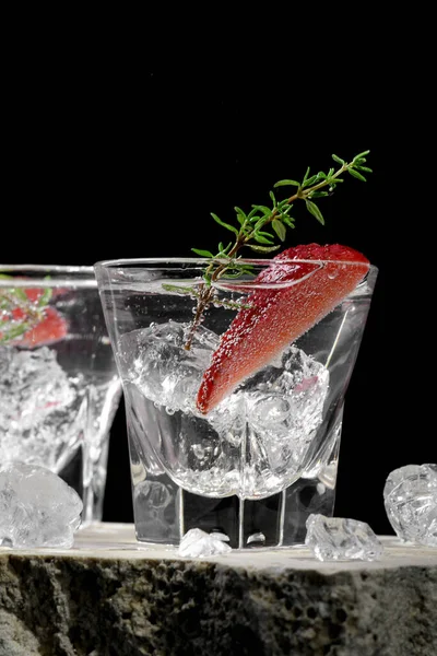 Vodka in two shot glass with ice, decorated with strawberry slice and thyme on travertine podium on dark stone background. Alcohol cocktail shot with strawberry and gin, fresh summer shots for party