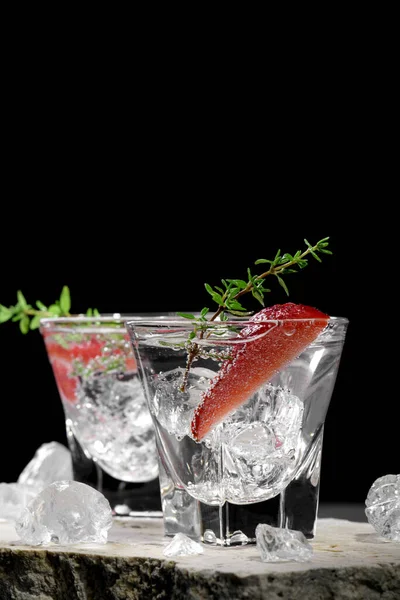 Vodka in two shot glass with ice, decorated with strawberry slice and thyme on travertine podium on dark stone background. Alcohol cocktail shot with strawberry and gin, fresh summer shots for party