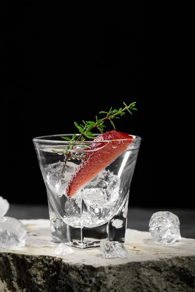 Vodka in shot glass with ice, decorated with strawberry slice and thyme on travertine podium on dark stone background. Alcohol cocktail shot with strawberry and gin, fresh summer shots for party