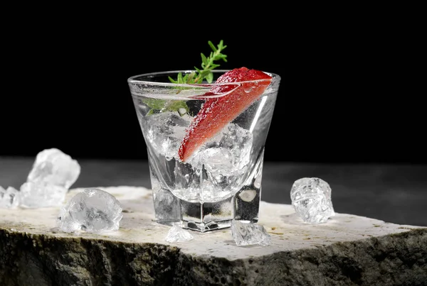 Vodka in shot glass with ice, decorated with strawberry slice and thyme on travertine podium on dark stone background. Alcohol cocktail shot with strawberry and gin, fresh summer shots for party