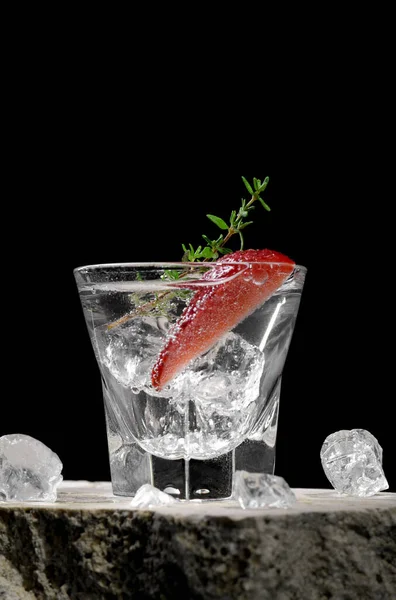 Vodka in shot glass with ice, decorated with strawberry slice and thyme on travertine podium on dark stone background. Alcohol cocktail shot with strawberry and gin, fresh summer shots for party