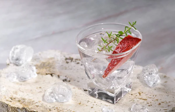 Vodka in shot glass with ice, decorated with strawberry slice and thyme on travertine podium on pink marble stone background. Alcohol cocktail shot with strawberry and vodka, fresh summer shots party