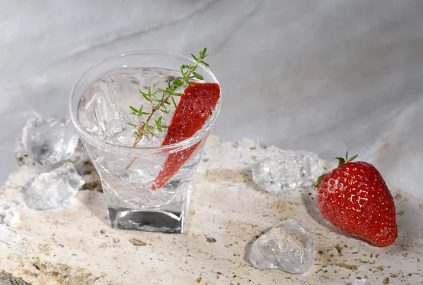 Vodka in shot glass with ice, decorated with strawberry slice and thyme on travertine podium on pink marble stone background. Alcohol cocktail shot with strawberry and vodka, fresh summer shots party