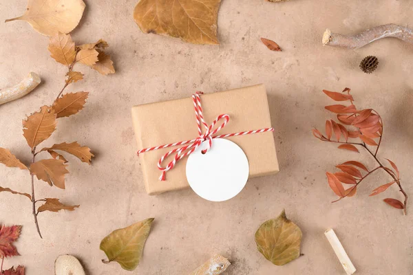 Autumn background: fallen leaves, dry plants with handmade gifts wrapped in craft paper and rope with gift tag mockup on natural beige background. Thanksgiving autumnal background, top view, flat lay