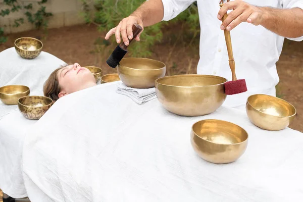 Young woman laying on a massage bed with tibetan singing bowl outdoor. A master of sound massage therapy doing a Buddhist healing practices, male hands holding a felt clappers