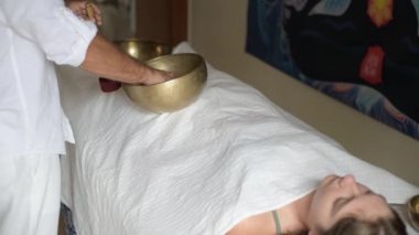 Beyazlı bir adam Tibet şarkı kasesiyle ses masajı yapıyor, Nepal Buddha şarkı kasesiyle ses terapisi yapıyor, genç bir kadın alternatif tıpla rahatlıyor.