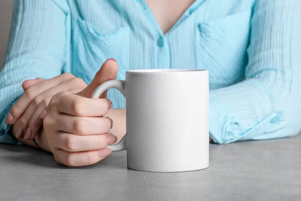 Female hand holding white mug with blank copy space for your advertising text message or promotional content. Girl in blue shirt holding white porcelain coffee mug mock up on the grey stone table