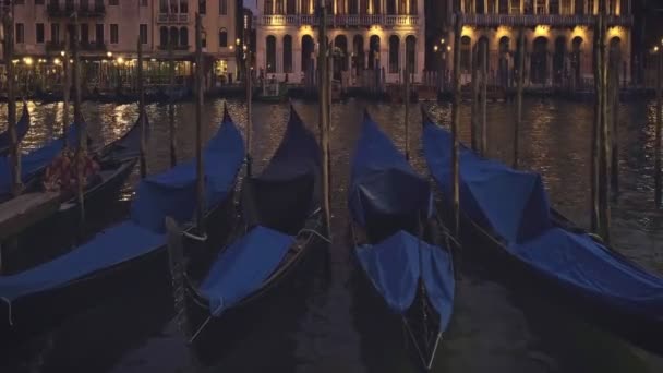 Gondels drijven 's nachts op het water van het Grand Canal in Venetië, Italië. Frontaal shot van geparkeerde boten met blauwe dekens, middeleeuwse gebouwen op de achtergrond — Stockvideo
