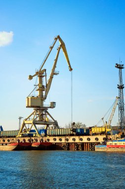 Güneşli bir günde nehir iskele vinci teleferikleri yüklüyor. Rıhtımdaki boş nehir sürükleme botları ya da mavnalar. Boş arabalar yüklemeye hazır. Dikey resim