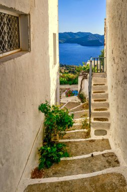 Deniz kenarındaki küçük Yunan köyünde günbatımında tipik manzara. Beyazlatılmış evler, saksılar, göl, asmalar. Geleneksel sokaklar, yaz güneşi, alçak güneş, mavi deniz suları, uzak tepeler, sessiz