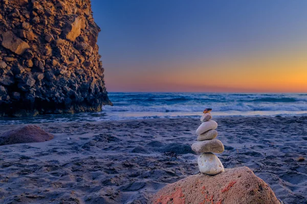 Small balance stone stack and big natural rock lit by sunset sun on sandy beach. Tidal waves in background, selective focus, blurred background, colorful view. Concept of balance, size, meditation