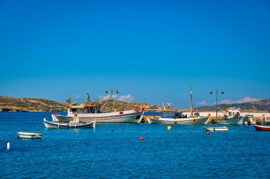 Yaz günü iskelede renkli balıkçı tekneleri ve Yunan ada köyünün tipik marinasında güneş ışığı. Akdeniz yaşam tarzı, tatiller, açık mavi gökyüzü ve uzak tepeler. Miloş, Kiklad, Yunanistan.