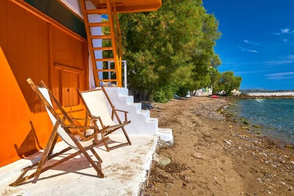 Two wooden beach chairs on porch of traditional Greek fisherman house just by sea tide. Whitewashed house walls and orange painted doors and stairs, no people, summer sunshine, sunny day, relax