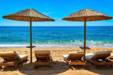 Gün ışığında kumlu sahilde turistler için iki güneş şemsiyesi ve dört güneşlik yatak hazır. Mükemmel bir yaz tatili. Mavi denizi ve kayalık tepeleri olan boş sahilde saman güneş gözlüğü. Ön çekim