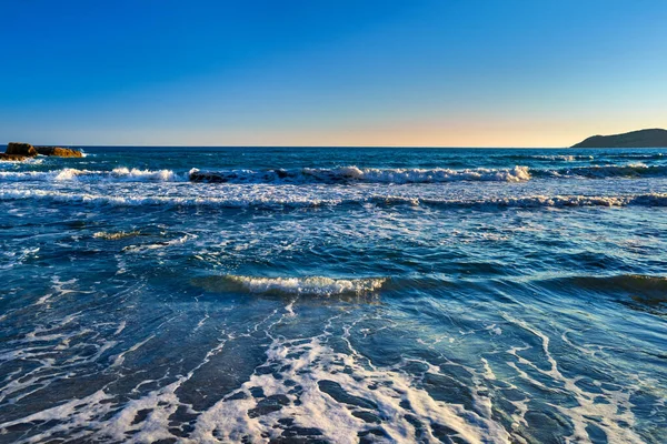 Beautiful seascape of low tidal waves, clear sunset sky, hazy hills. Clear blue and azure waters approaching with little white foam. Nature, landscape, calm and relaxing meditative view. Mediterranean