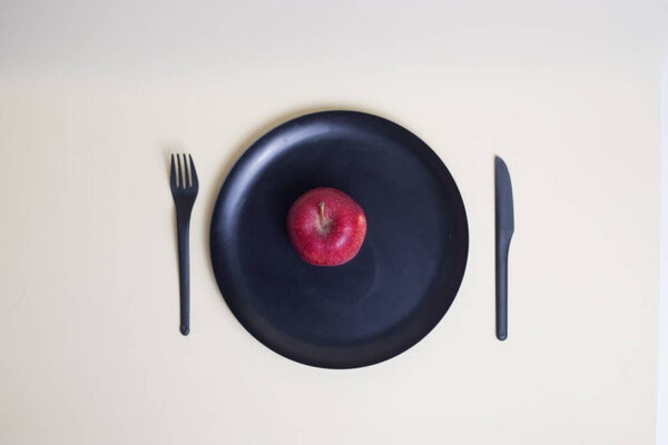 red apple on black plate with black plastic knife and fork isolated on light background flat lay. Image contains copy space