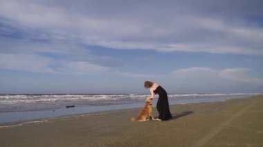 Uzun siyah elbiseli mutlu genç kadın dışarıda labrador köpeğiyle sahilde yürüyor.