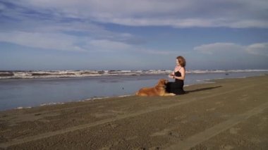 Uzun siyah elbiseli mutlu genç kadın dışarıda labrador köpeğiyle sahilde yürüyor.