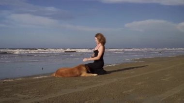 Uzun siyah elbiseli mutlu genç kadın dışarıda labrador köpeğiyle sahilde yürüyor.