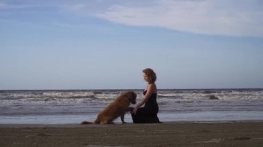 Uzun siyah elbiseli mutlu genç kadın dışarıda labrador köpeğiyle sahilde yürüyor.