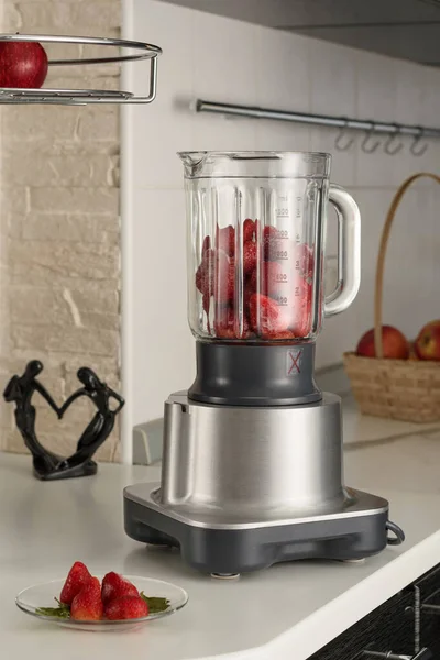Smoothie yapmak için çilekli bir blender, arka planda mutfak masasının üzerinde duruyor. Tabaktaki biblo ve çileklerin yanında. Arka planda elma dolu bir sepet var..