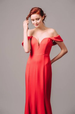 Kırmızı uzun elbiseli, yüksek topuklu yakalı, parlak genç bir bayan gri stüdyo arka planında yürüyor ve gülümsüyor. Şenlikli kadın elbisesi. 