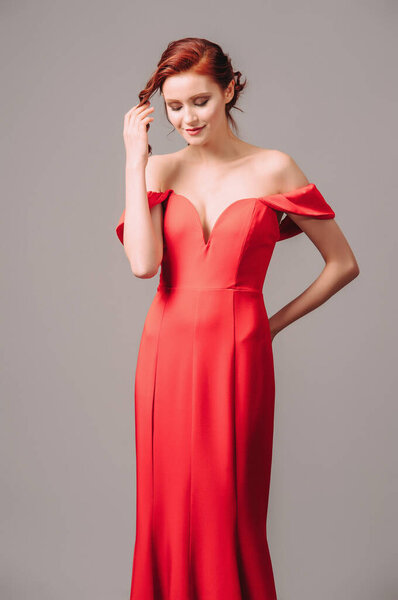 Светлая юная леди в алом длинном платье с глубоким вырезом на высоких каблуках, идущая и улыбающаяся на сером фоне студии. Праздничное женское платье. 