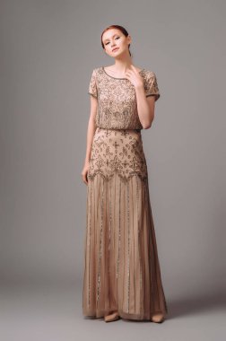 Zarif bej üzerine uzun bir elbise. Kızıl kadın stüdyoda poz veriyor. Eski tarz, kısa kollu ve kahverengi boncuklu bir gece elbisesi. Kadın cazibesi kıyafeti..