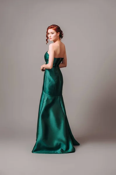 Emerald ball gown. Young lady in long green evening dress. Ginger woman posing in studio. Beautiful model wearing high heels, feminine look for an event. Women\'s classic fashion.