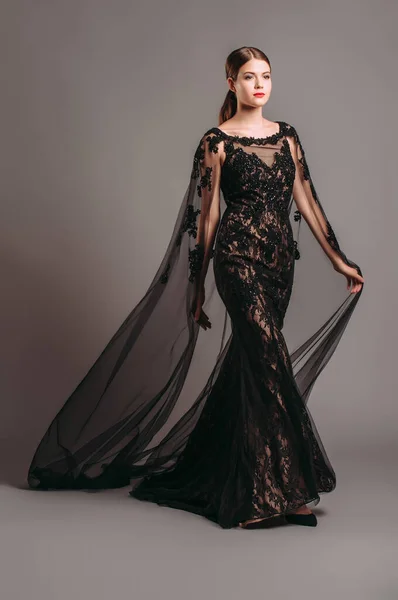 Şeffaf pelerinli, siyah, dantelli bir gece elbisesi. Trenle nakışlı Maxi elbise, modern moda. Balıkkuyruklu elbise. Esmer model stüdyoda poz veriyor. Tamamen siyah kadın görünüşü.. 