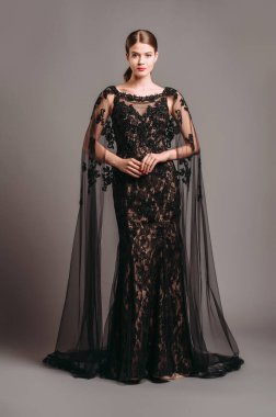Şeffaf pelerinli, siyah, dantelli bir gece elbisesi. Trenle nakışlı Maxi elbise, modern moda. Balıkkuyruklu elbise. Esmer model stüdyoda poz veriyor. Tamamen siyah kadın görünüşü.. 