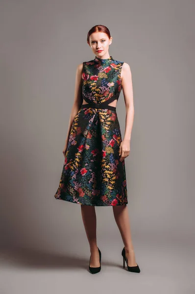 Cut out waist midi dress in floral embroidery with black high heels. Ginger lady posing in studio. Evening colourful elegant gown with high neck line, female fashion, gorgeous chic look.