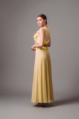Sleeveless evening gown with fluttering pleating skirt. Long yellow summer dress, rear view. Beautiful young lady posing in studio. Fashionable feminine look. 