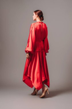 Uzun kollu, uzun kollu ve yüksek topuklu seksi yakalı, gri stüdyo arka planında mırıldanan kırmızı büyük beden elbiseli parlak genç bayan. Zarif çağdaş kadın görünüşü. 