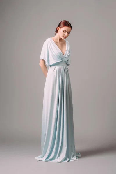 Full-length blue bridesmaid dress with flutter sleeves. Effortless festive summer look. Ginger lady in elegant, romantic, tender evening gown. Studio portrait of a young smiling woman.
